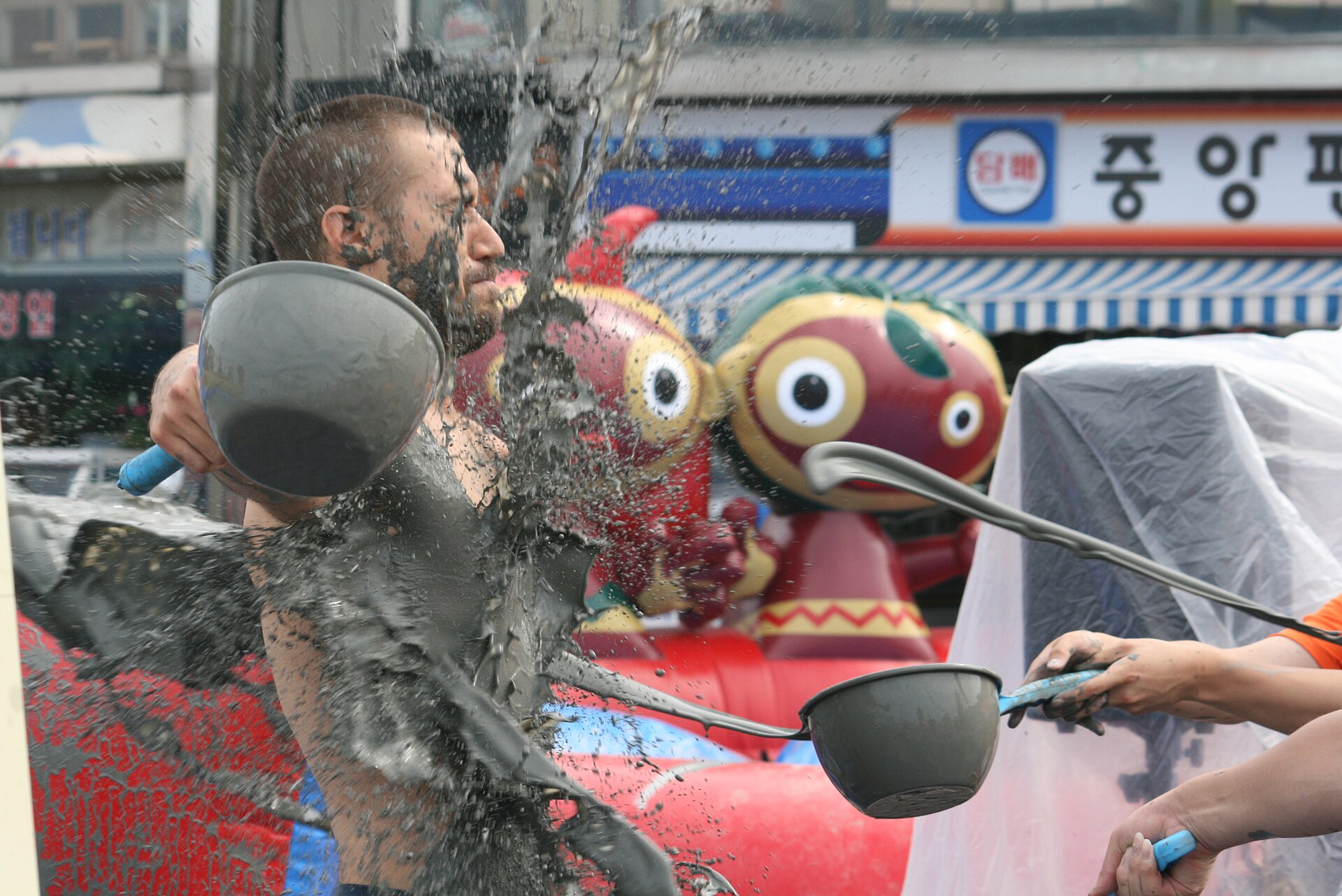 A festival-goer getting splashed with grey mud at the Boryeong Mud Festival, with the festival's mud mascots behind