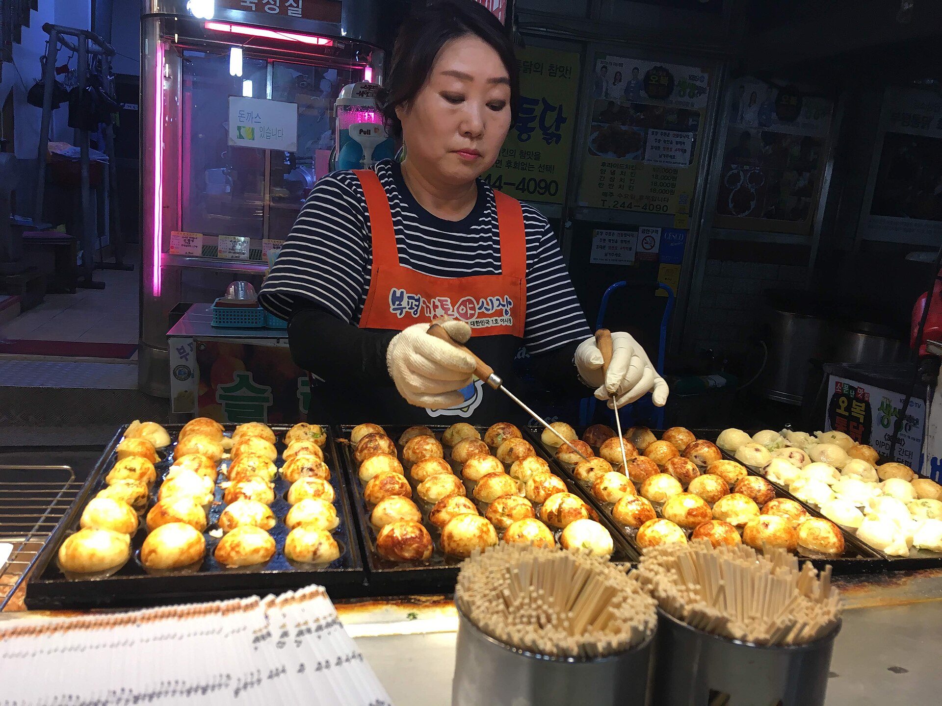A vendor at Bupyeong Kkangtong Night Market in Busan turning takoyaki on a hot plate, her apron reading ‘Bupyeong Kkangtong Night Market — Korea’s first night market’