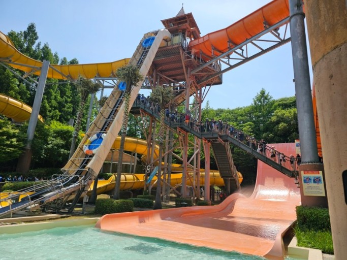 The fortress-style water-slide tower at Caribbean Bay in Yongin, with orange and yellow slides winding down to a turquoise splash pool, framed by palm trees under a bright summer sky.