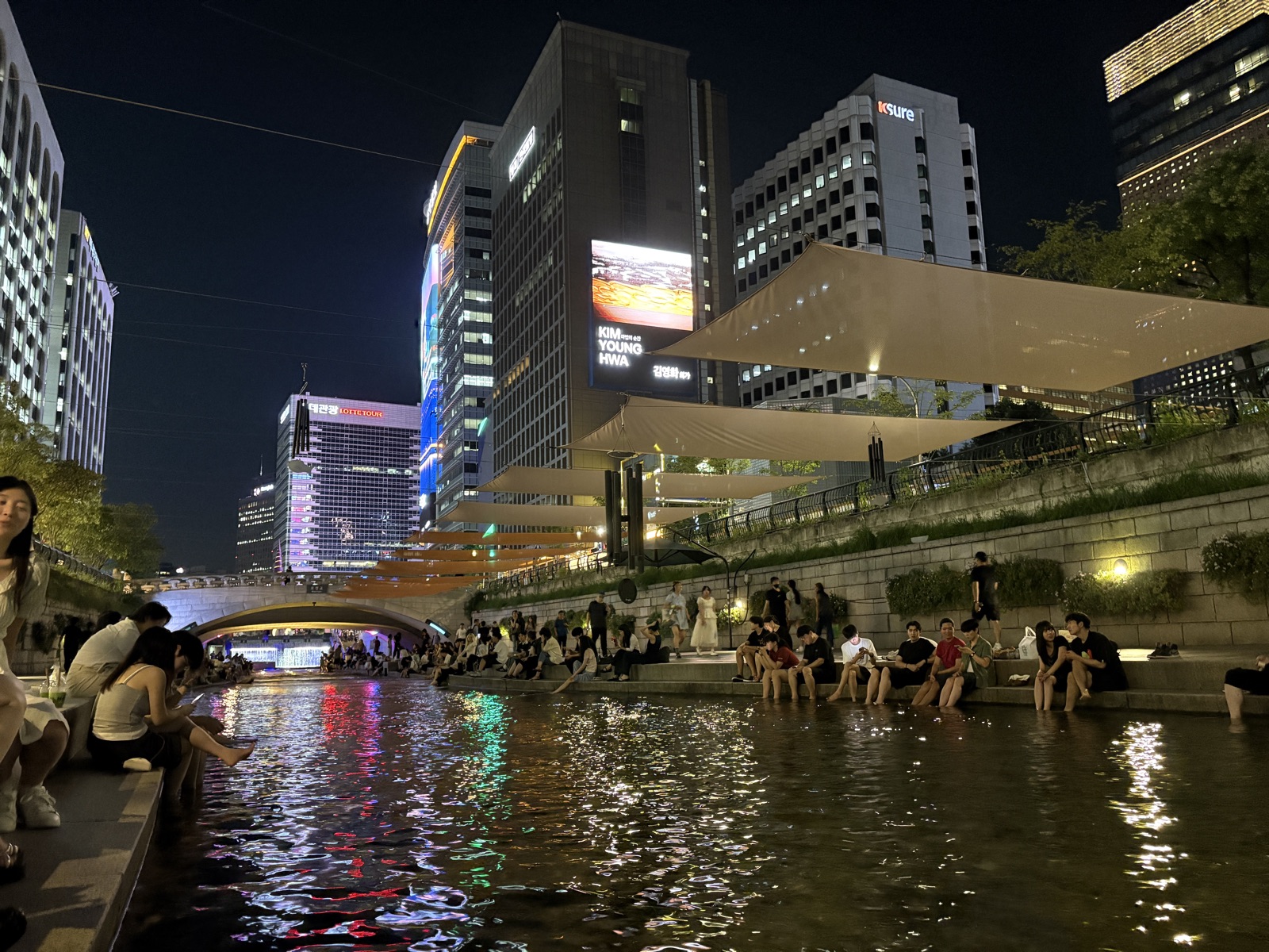 People sitting along Cheonggyecheon stream on a summer evening in Seoul, cooling their feet in the water with the city lights reflected