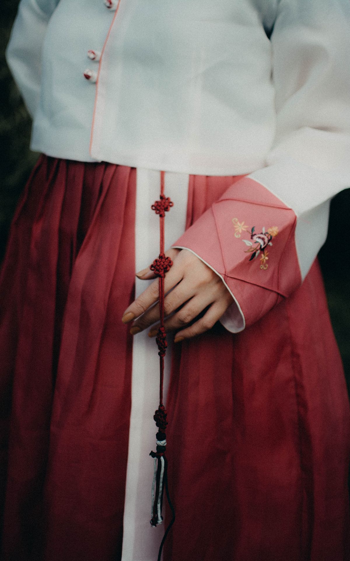 A person wearing a white and deep-red hanbok, with a decorative knotted norigae tassel hanging from the waist.