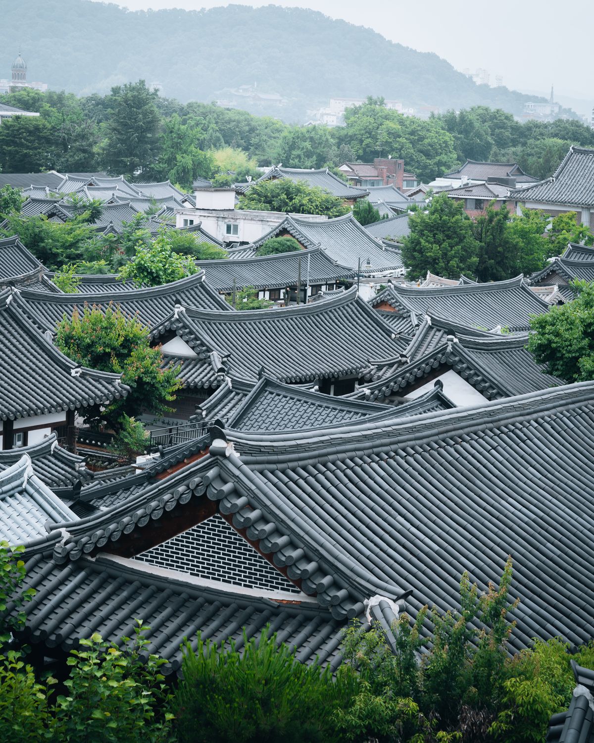Layered grey-tiled curved roofs of a traditional Korean hanok village, with green trees and a misty hill behind.