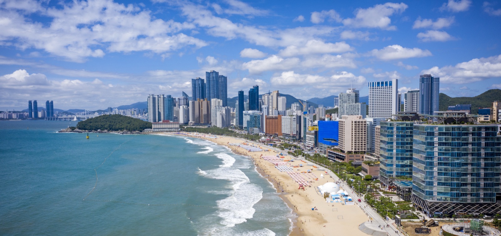 Aerial view of Haeundae Beach in summer — rows of parasols on the sand, waves rolling in, and the high-rise skyline behind