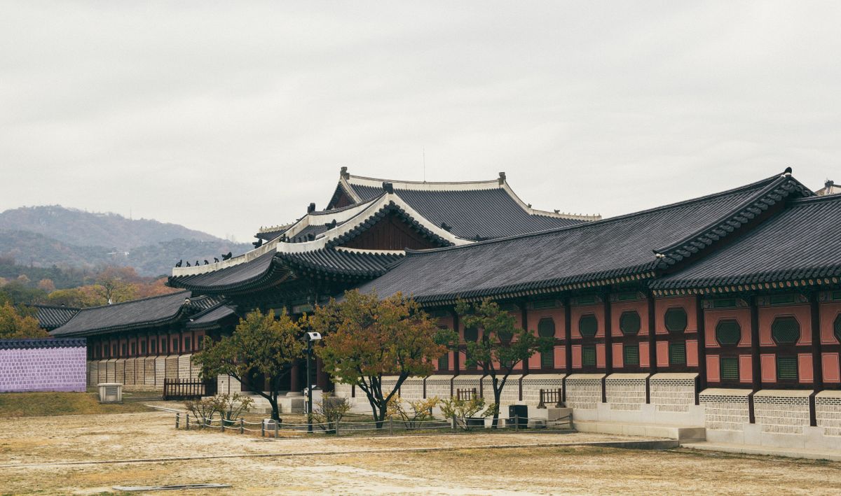 A long Korean royal palace hall with sweeping black-tiled roofs and red-and-green painted eaves, set on a wide gravel courtyard with low autumn-tinged hills behind.